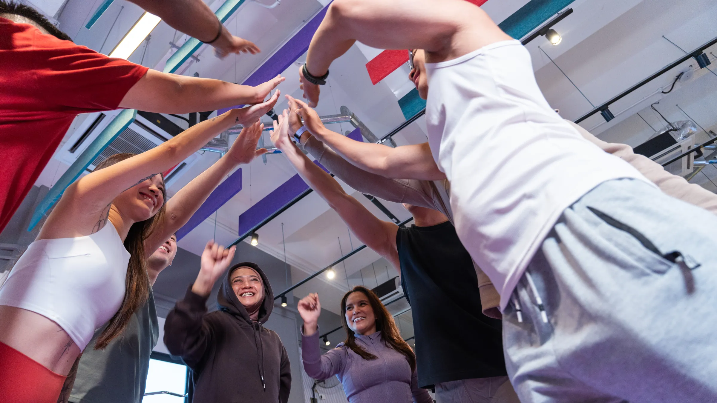Fitness group in workout clothes joining hands in a cheer inside a gym with colorful ceiling panels.
