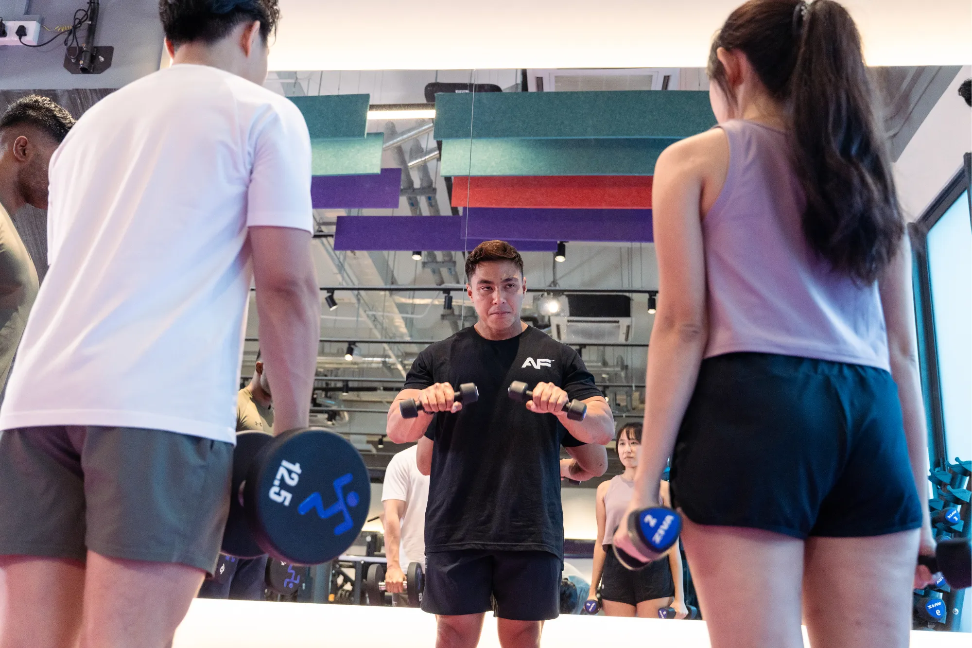 A man and a woman lifting dumbbells in a gym in front of a mirror with colourful acoustic panels overhead.