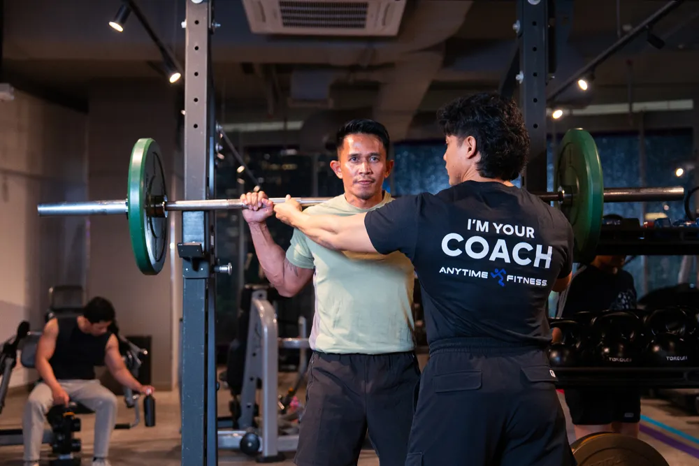 Pelatih kebugaran memperhatikan seorang pria yang mengangkat barbel di gym dengan beban dan peralatan di sekelilingnya.