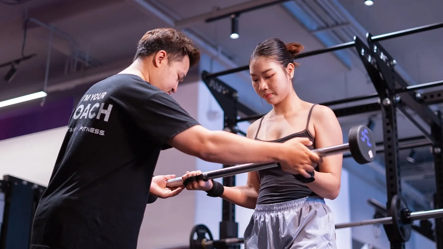 เทรนเนอร์ Anytime Fitness กำลังช่วยจัดท่าจับบาร์เบลให้สมาชิกหญิง