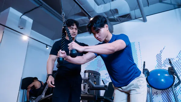 Trainer coaching man using cable machine for strength exercise at a gym.