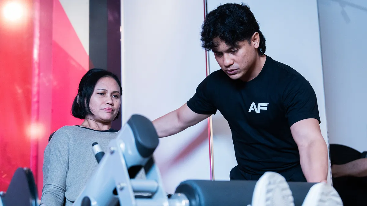 Man in black shirt assisting woman exercising on gym leg press machine.