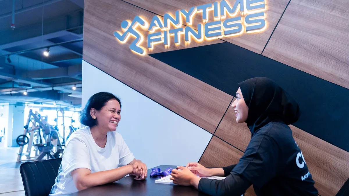 Two women smiling and talking at a table inside an Anytime Fitness gym lobby.