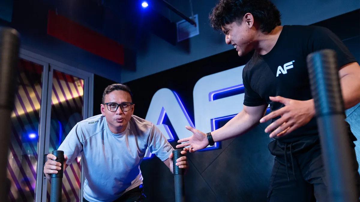 Man in glasses exercises with gym trainer spotting him beside a wall with lit AF logo.