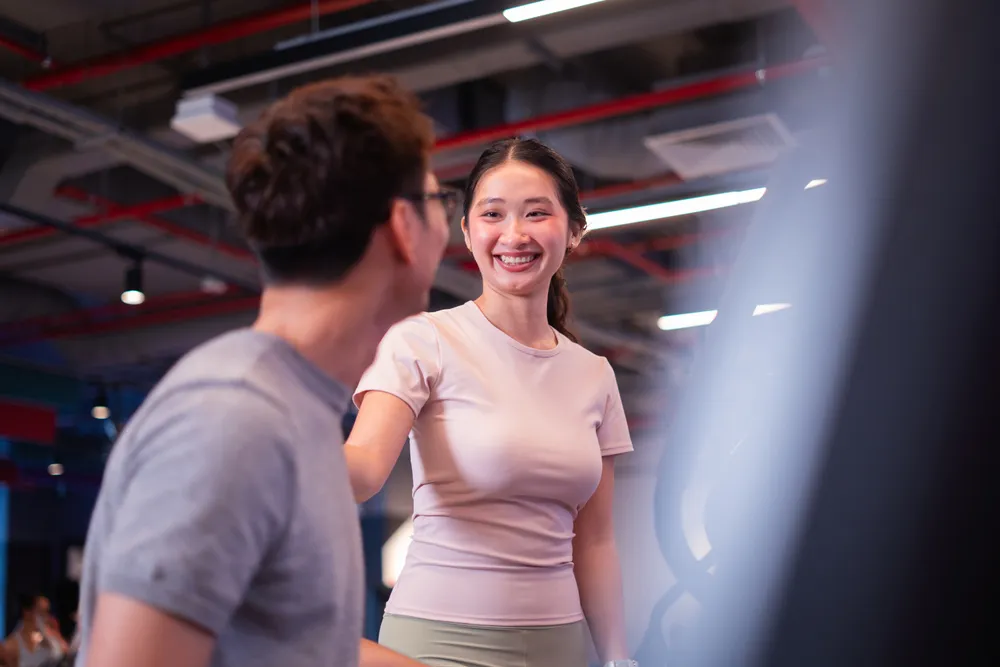 Người phụ nữ mỉm cười mặc áo hồng đang trò chuyện với một người đàn ông trong bối cảnh phòng tập, với đèn trần công nghiệp.
