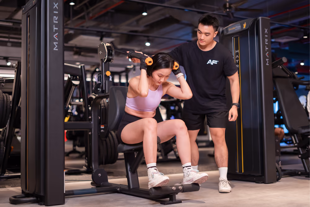 Người phụ nữ đang thực hiện bài tập gập bụng trên máy ghế tập gym với huấn luyện viên nam hướng dẫn cô ấy.