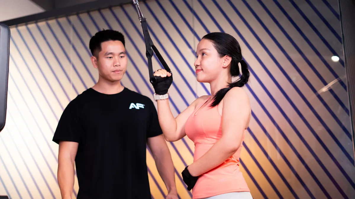 Người phụ nữ mặc áo ba lỗ màu hồng đang tập với dây kháng lực dưới sự giám sát của huấn luyện viên phòng tập.