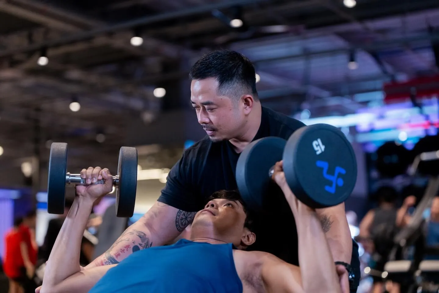 Personal trainer spotting a man performing a dumbbell chest press on a weight bench.