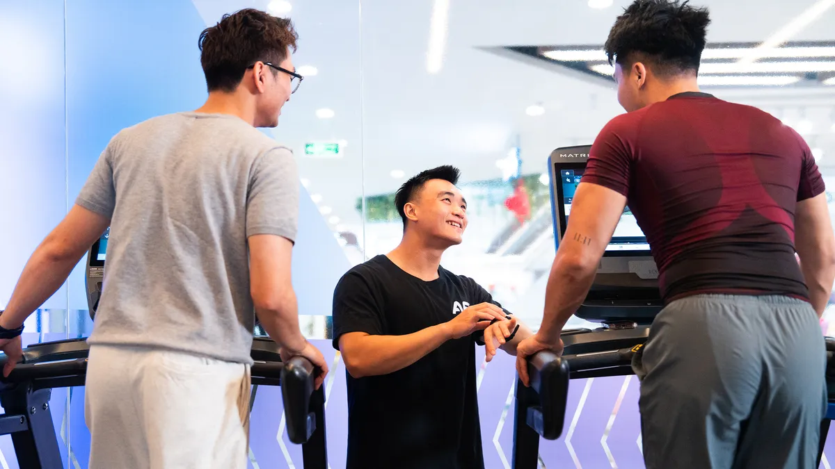 Huấn luyện viên nói chuyện với hai người đàn ông trên máy chạy bộ trong môi trường phòng tập thể dục hiện đại.