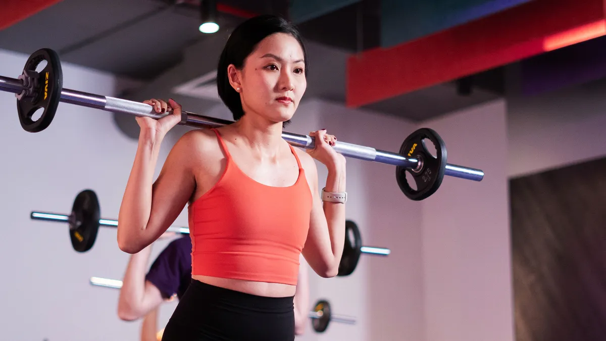 Phụ nữ mặc áo màu cam đang nâng tạ đòn trên vai tại một lớp tập luyện ở phòng gym.