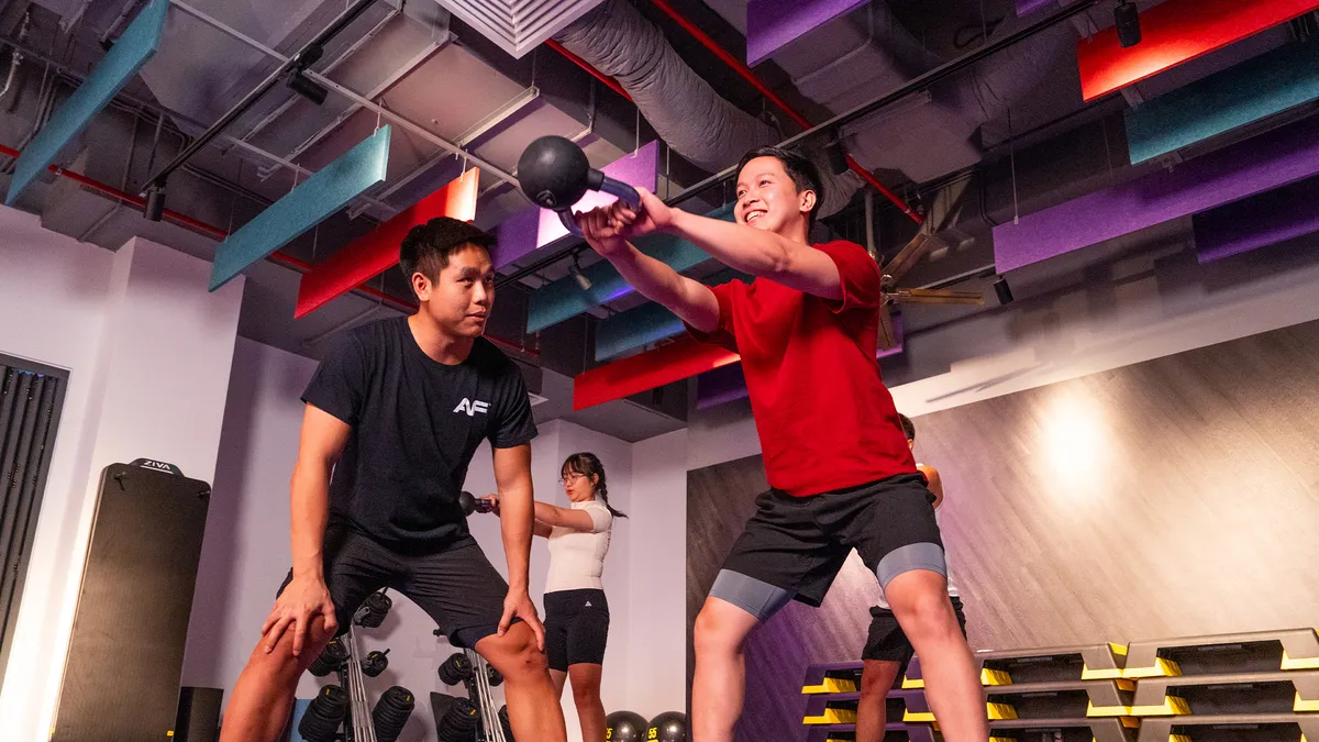 Người phụ nữ mặc áo đỏ đang vung tạ ấm trong buổi tập thể dục cùng huấn luyện viên nam trong phòng gym.