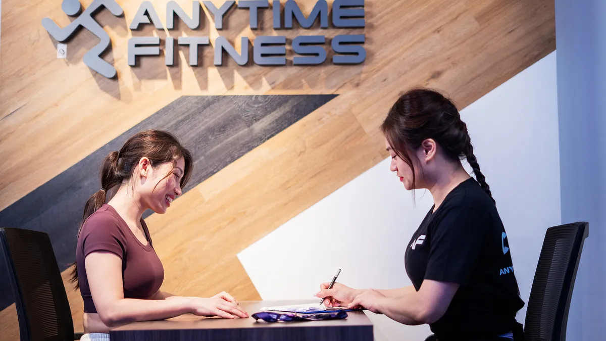 Hai phụ nữ ngồi tại bàn trong một phòng tập Anytime Fitness, một người mỉm cười còn người kia đang viết trên bảng kẹp giấy.
