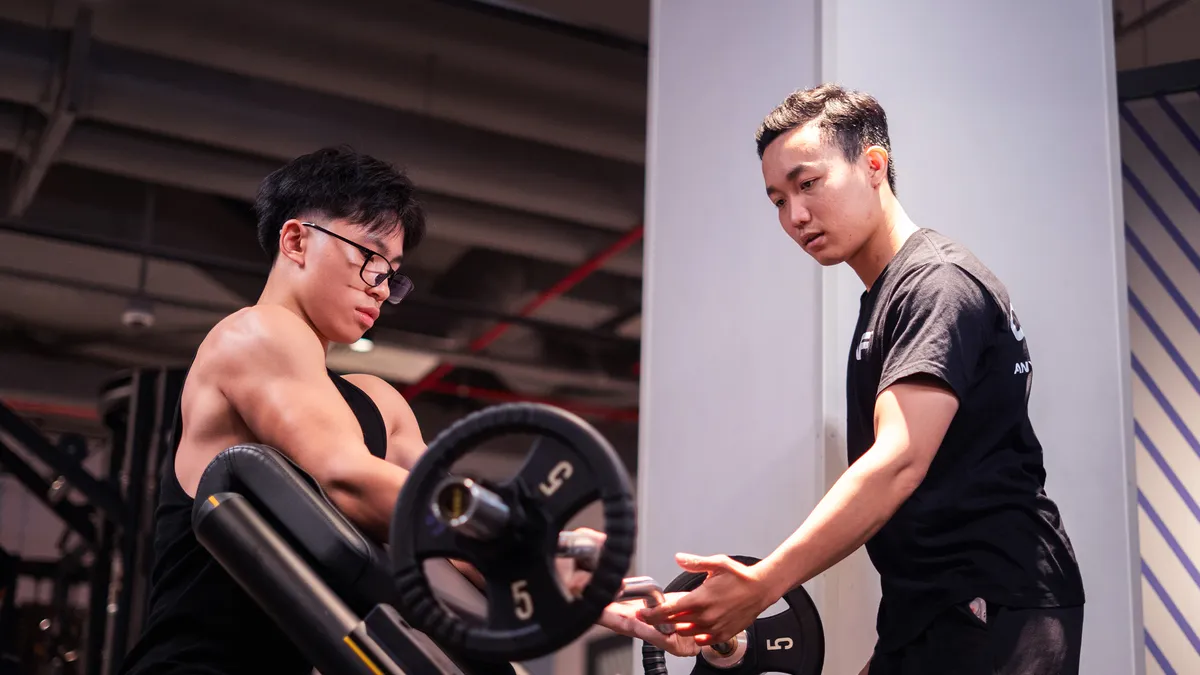 Một người đàn ông mặc áo sơ mi đen đang hỗ trợ một người đàn ông nâng tạ tại phòng tập.