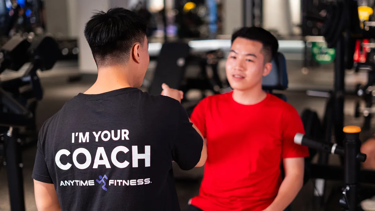 Huấn luyện viên thể hình mặc áo "I'm your coach" đang nói chuyện với một chàng trai trẻ mặc áo đỏ tại phòng gym.