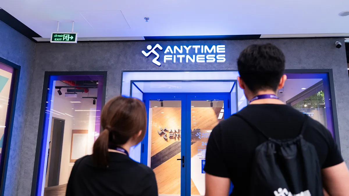 Hai người từ phía sau đang nhìn vào lối vào của một phòng gym Anytime Fitness với cửa màu xanh và biển hiệu sáng đèn.