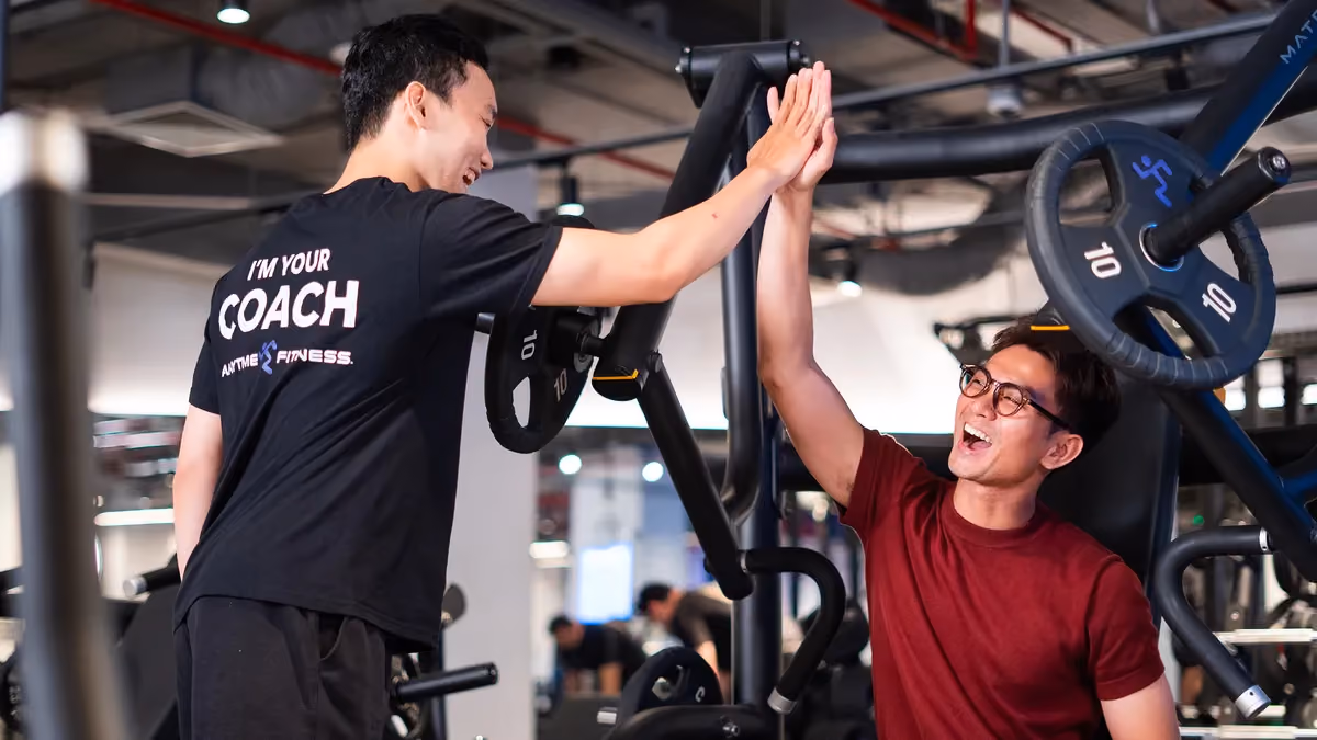Gym coach and man in glasses high-five each other near weightlifting machine.