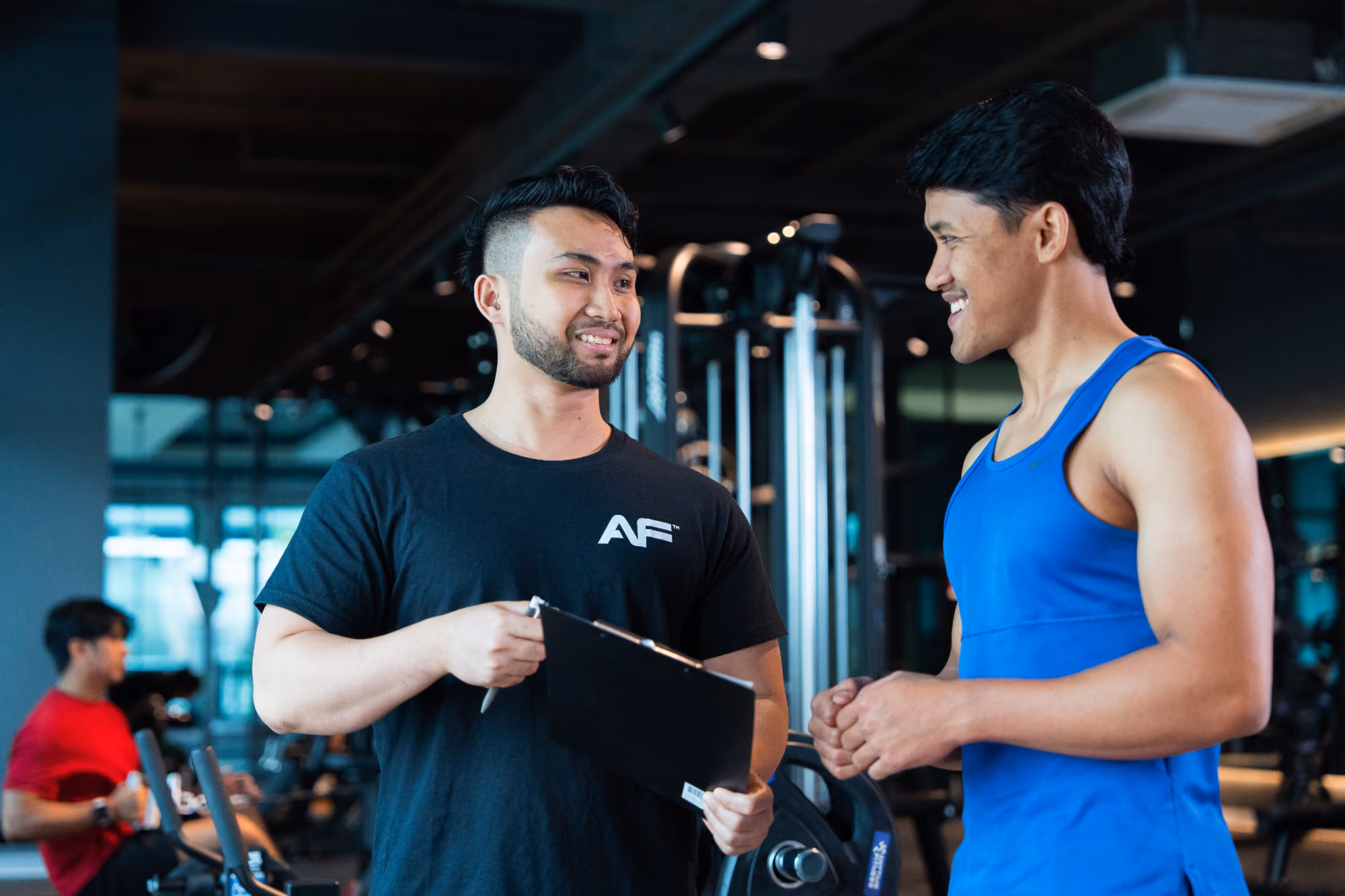 Two men in gym having a friendly conversation, one holds a clipboard, workout equipment visible.