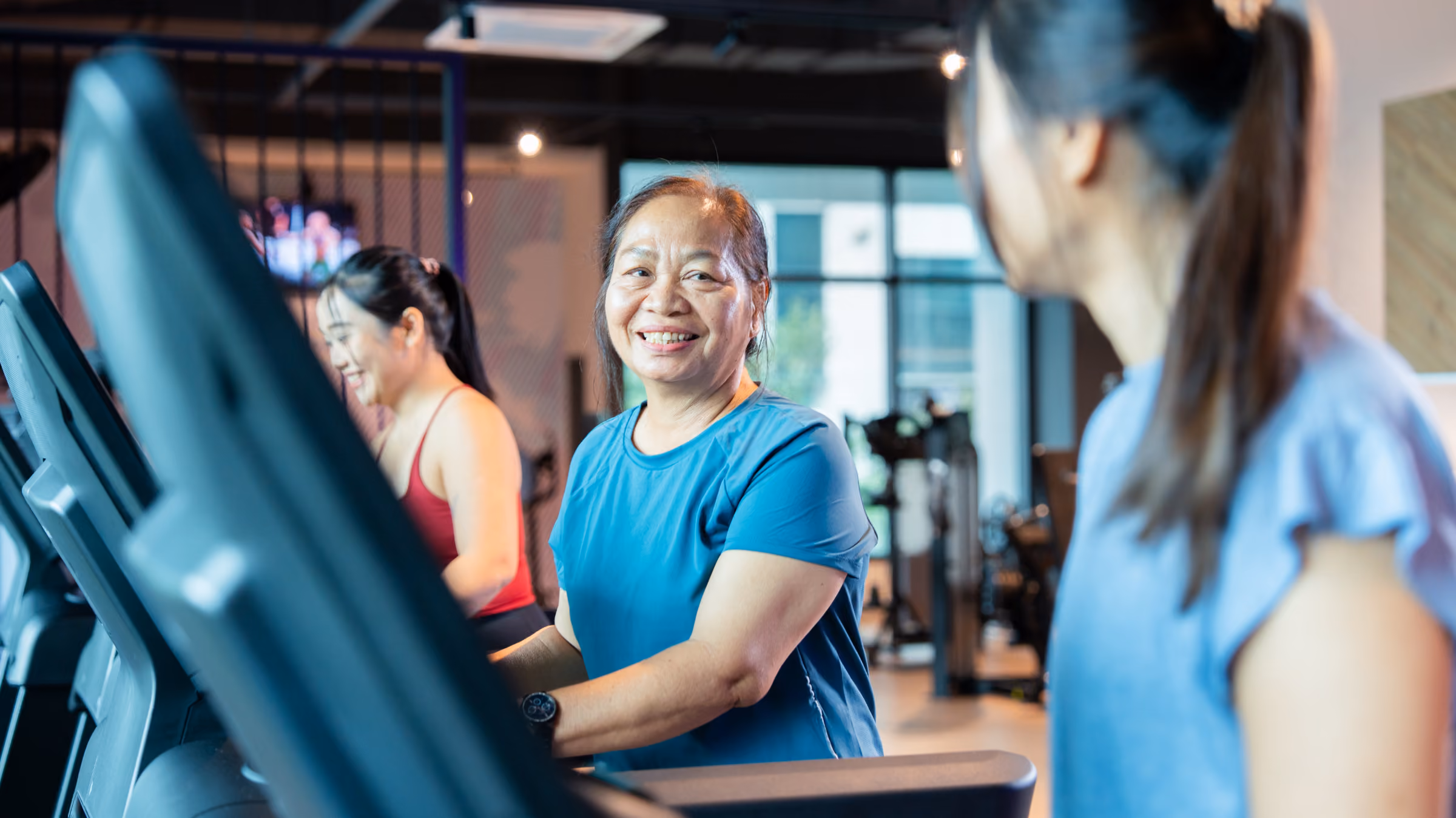 Three women exercising on treadmills in a gym, one smiling and looking at another.