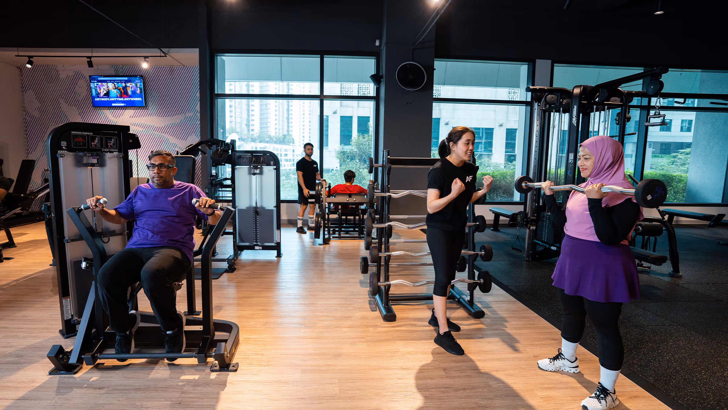 People exercising on gym machines and a woman lifting weights with trainer encouragement visible.