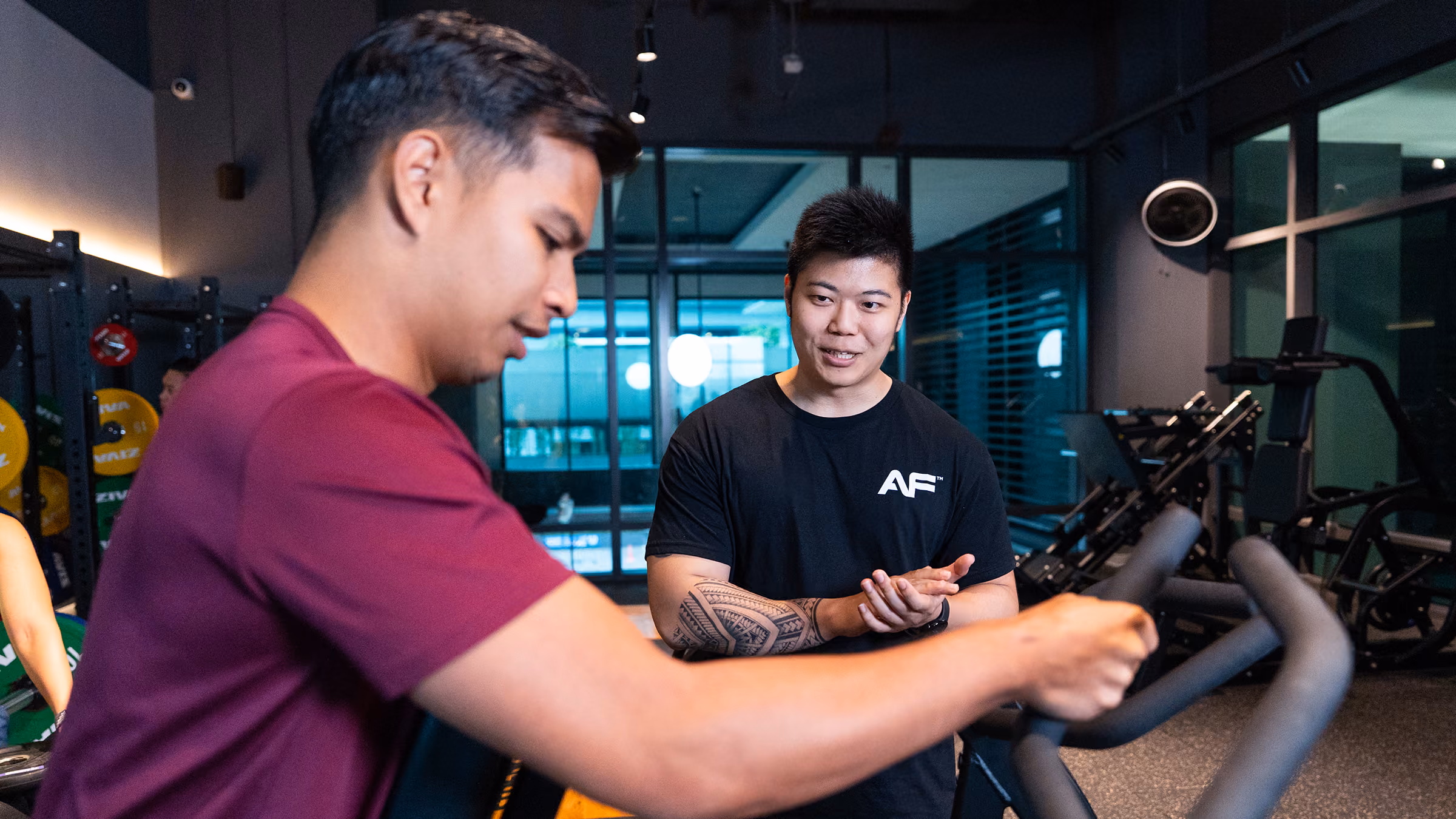 Trainer with tattoo coaching man in maroon shirt using gym exercise equipment indoors.