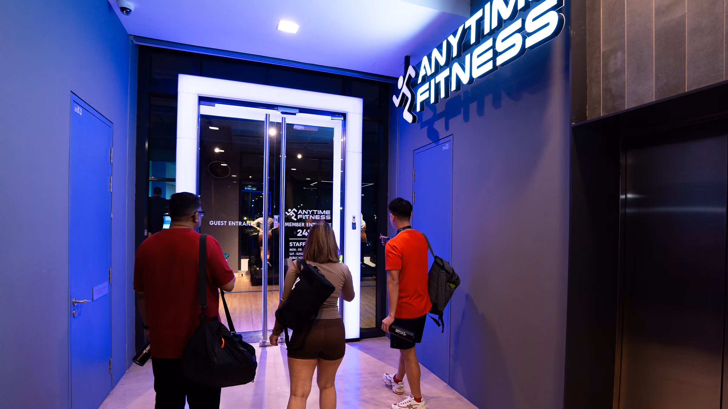 Three people with bags enter an Anytime Fitness gym through glass doors with bright lights.