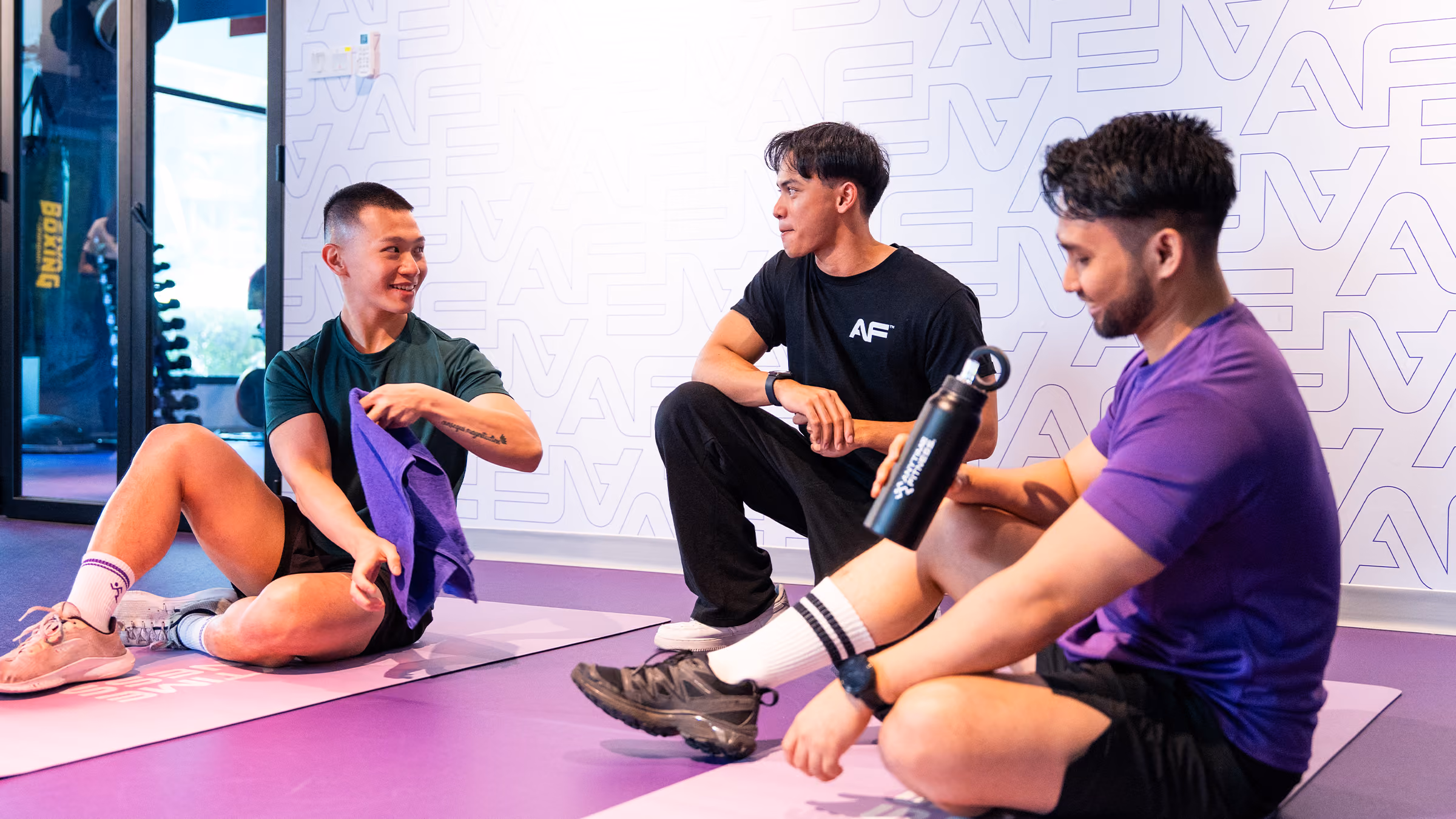 Three men resting on exercise mats in a gym, one holding a towel, another holding a water bottle.