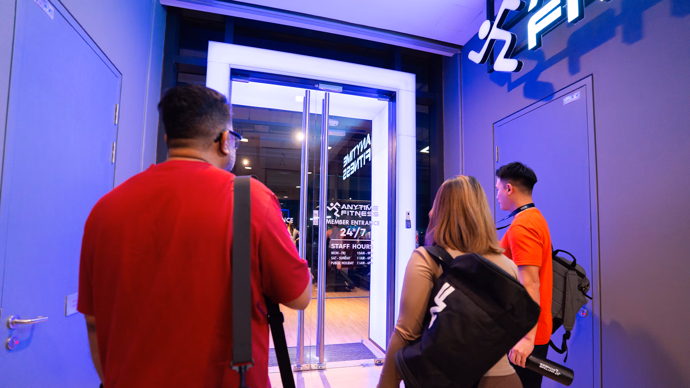 Three people entering an Anytime Fitness gym through glass doors with visible club hours sign.