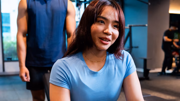 Woman in blue shirt smiling and seated in a gym with a man standing behind her.