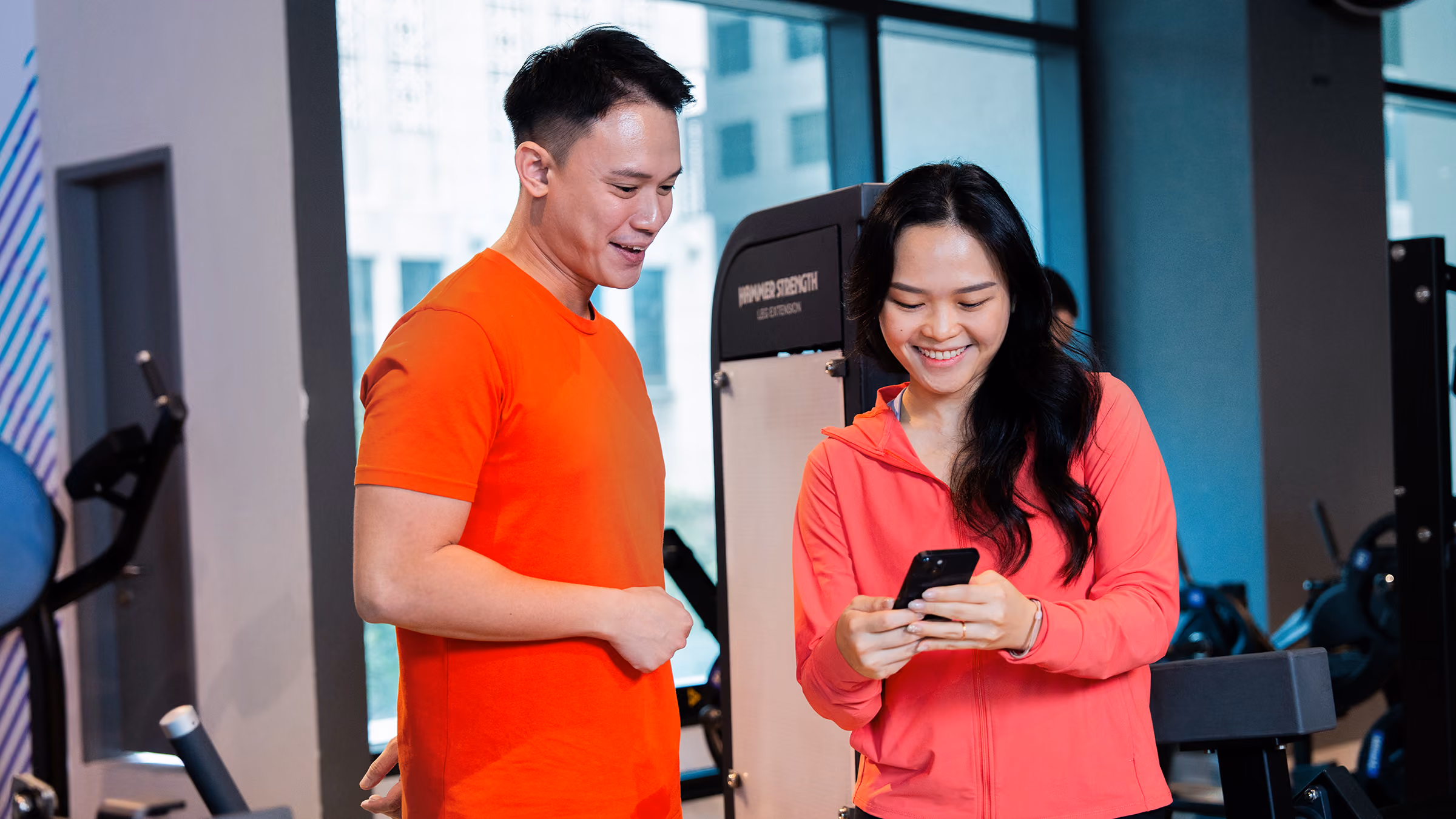 Man and woman in a gym looking at a phone together, smiling and wearing bright workout clothes.