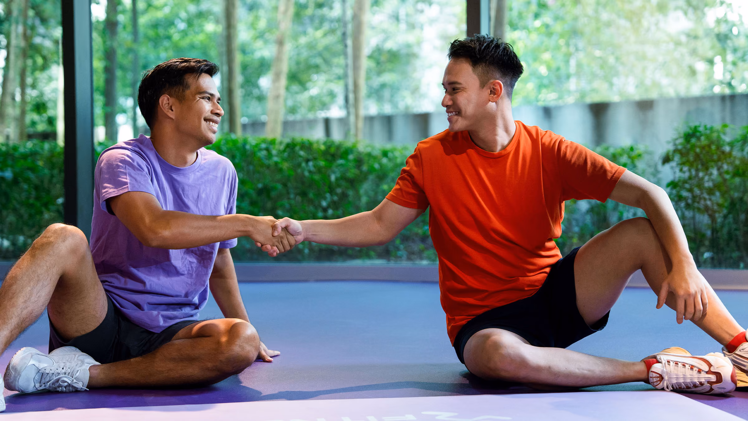 Two men sitting on a gym floor, facing each other and shaking hands with smiles.