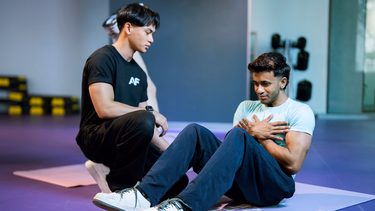 A personal trainer assists a man lifting heavy dumbbells on a bench in a busy gym with various equipment in the background.