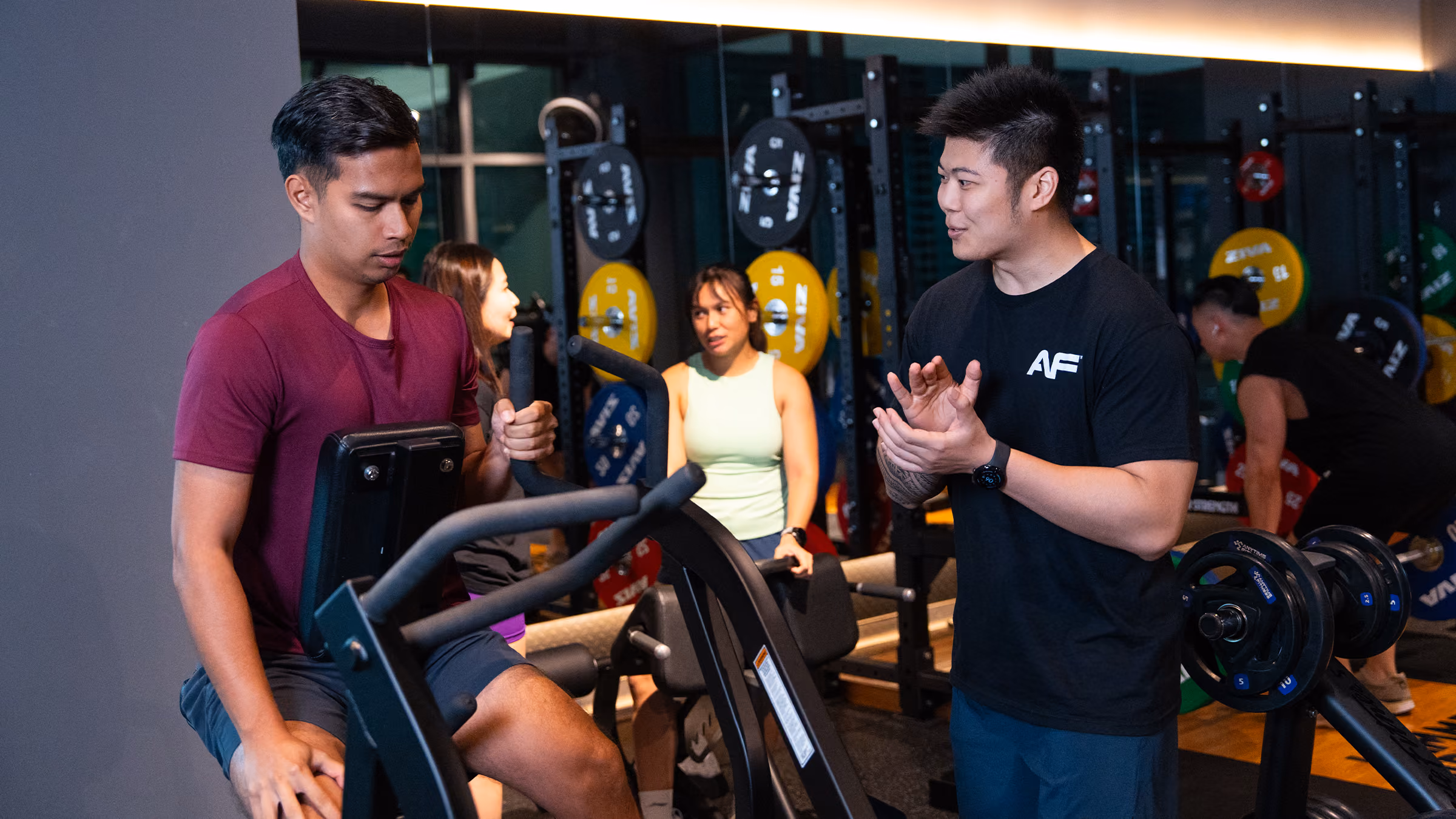 Man in maroon shirt exercises on a machine while a trainer in black coaches him in a gym.