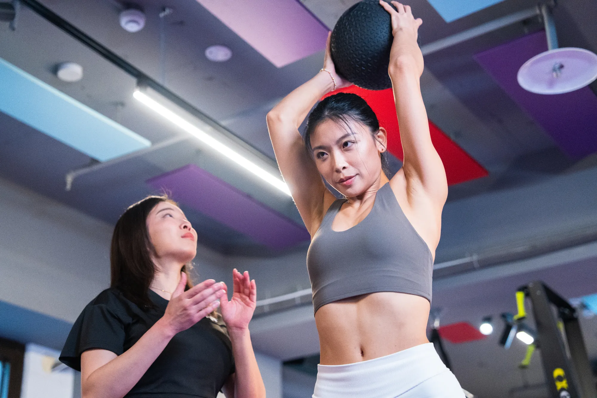 女人在頭上舉重的球，而另一位女人在健身房看並打手。