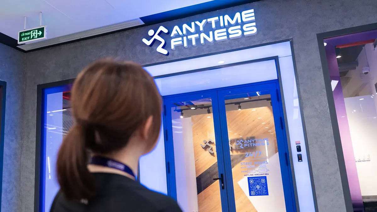 Người nhìn vào lối vào của phòng tập thể dục Anytime Fitness với cửa màu xanh và bảng hiệu thắp sáng.