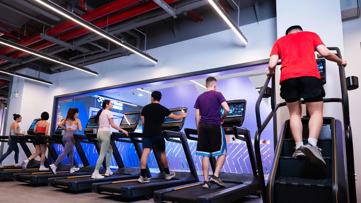 Sáu người tập thể dục trên máy chạy bộ và máy leo cầu thang trong một phòng tập thể dục hiện đại.