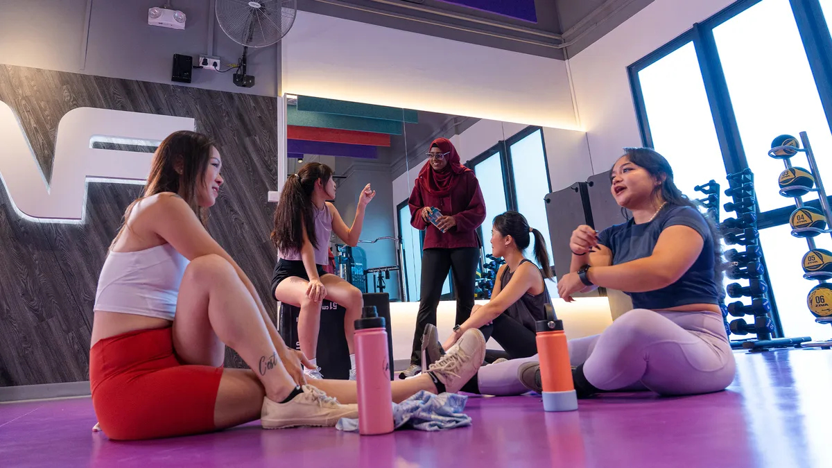 Five diverse women resting and chatting in a gym with water bottles and exercise equipment nearby.