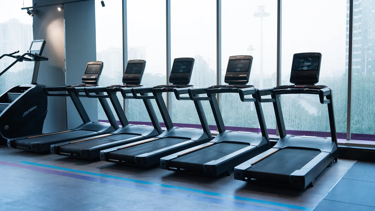 Row of six black treadmills lined up in a gym near large windows with a city view.