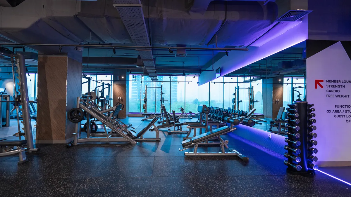 Modern gym interior with weight machines, dumbbells, and large windows showing city buildings.