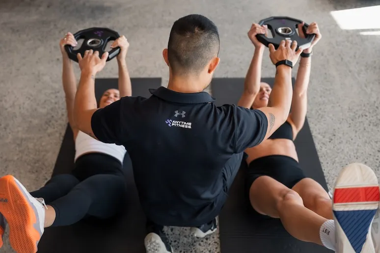 Fitness trainer guides two women holding weights while lying on mats exercising legs in a gym.