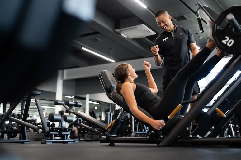 Trainer encouraging woman using leg press machine in a gym with fitness equipment around.