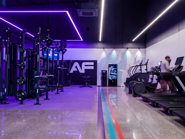 Modern gym interior with weightlifting area, treadmills, neon lights, and a person using a treadmill.