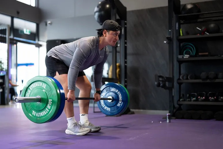 Man in gray shirt and cap lifting barbell with green and blue weights in gym with purple floor.
