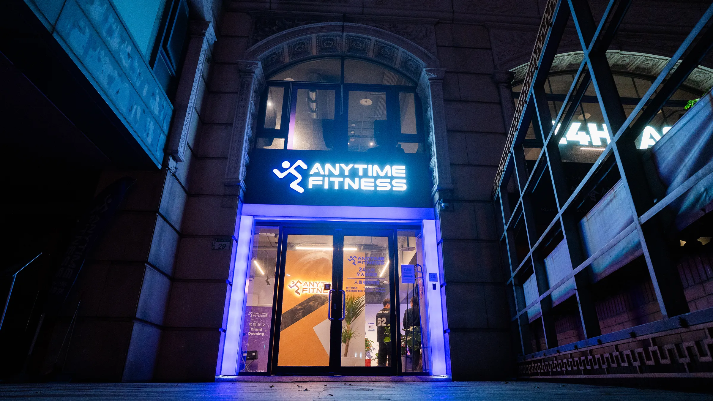 Anytime Fitness 健身房的入口，晚上以藍燈點亮，室內有兩人。