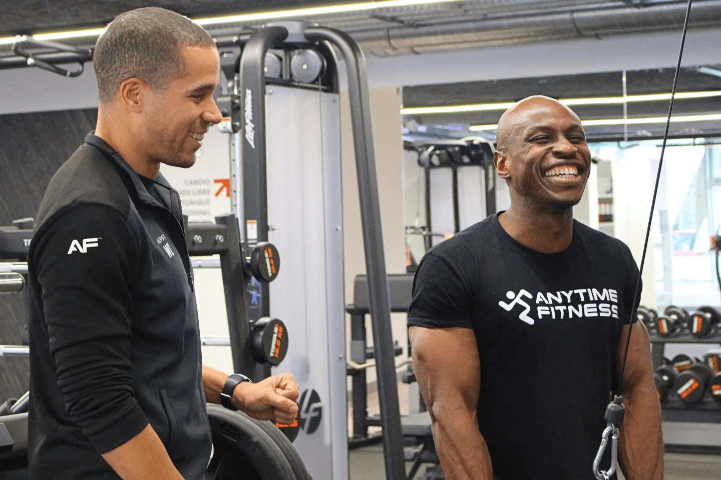 Un membre d'Anytime Fitness souriant pendant son entraînement avec une poulie, encouragé par un coach personnel.