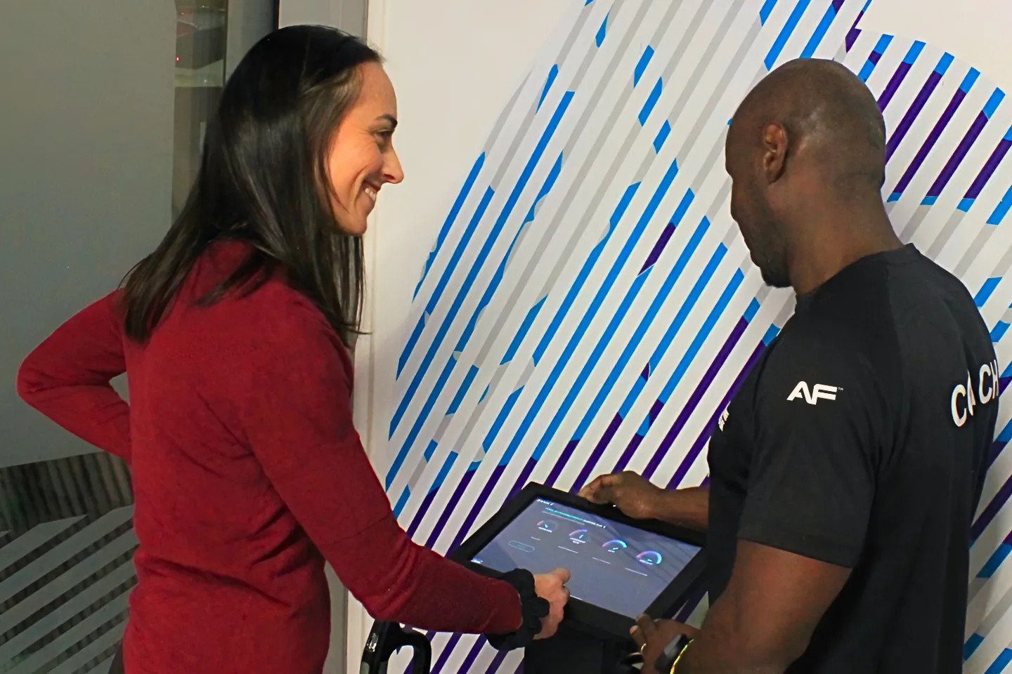 Une membre souriante d'Anytime Fitness échangeant avec un coach personnel devant un écran tactile d'évaluation fitness.