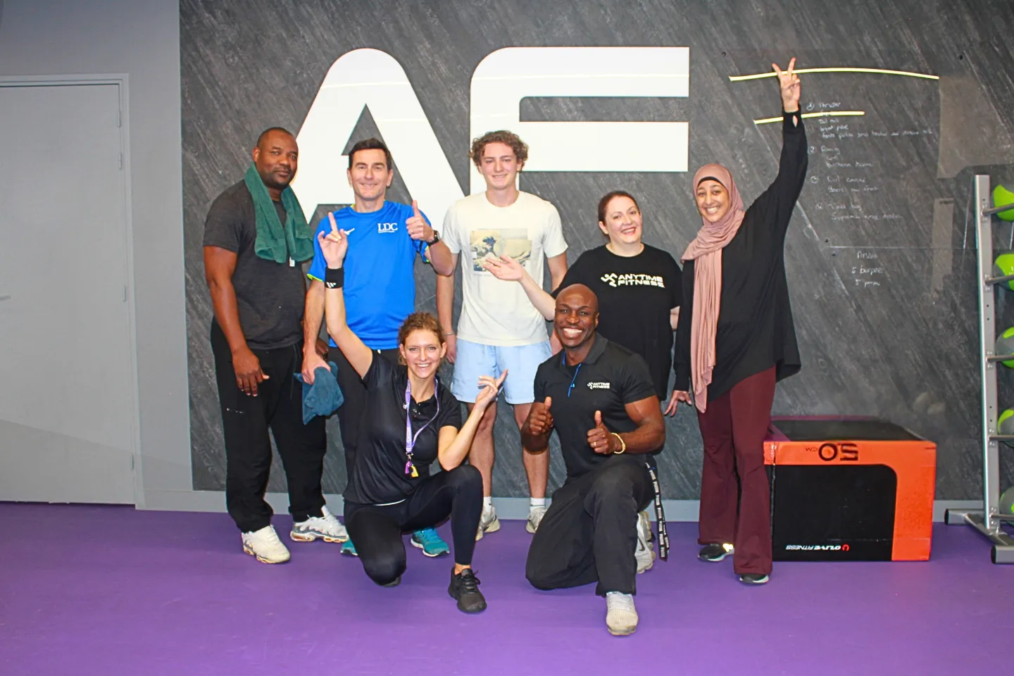 Un groupe diversifié de membres et de coachs Anytime Fitness posant joyeusement ensemble dans la salle de sport devant le logo AF.