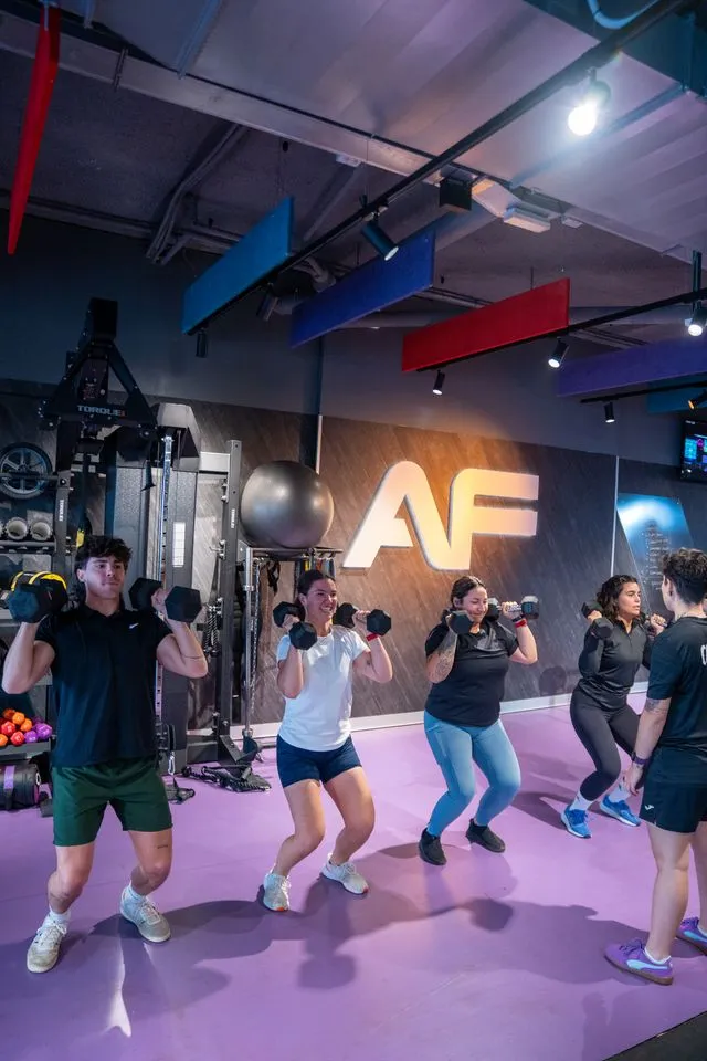 Grupo de personas en un gimnasio haciendo ejercicios de prensa de hombros con mancuernas sobre un piso morado.