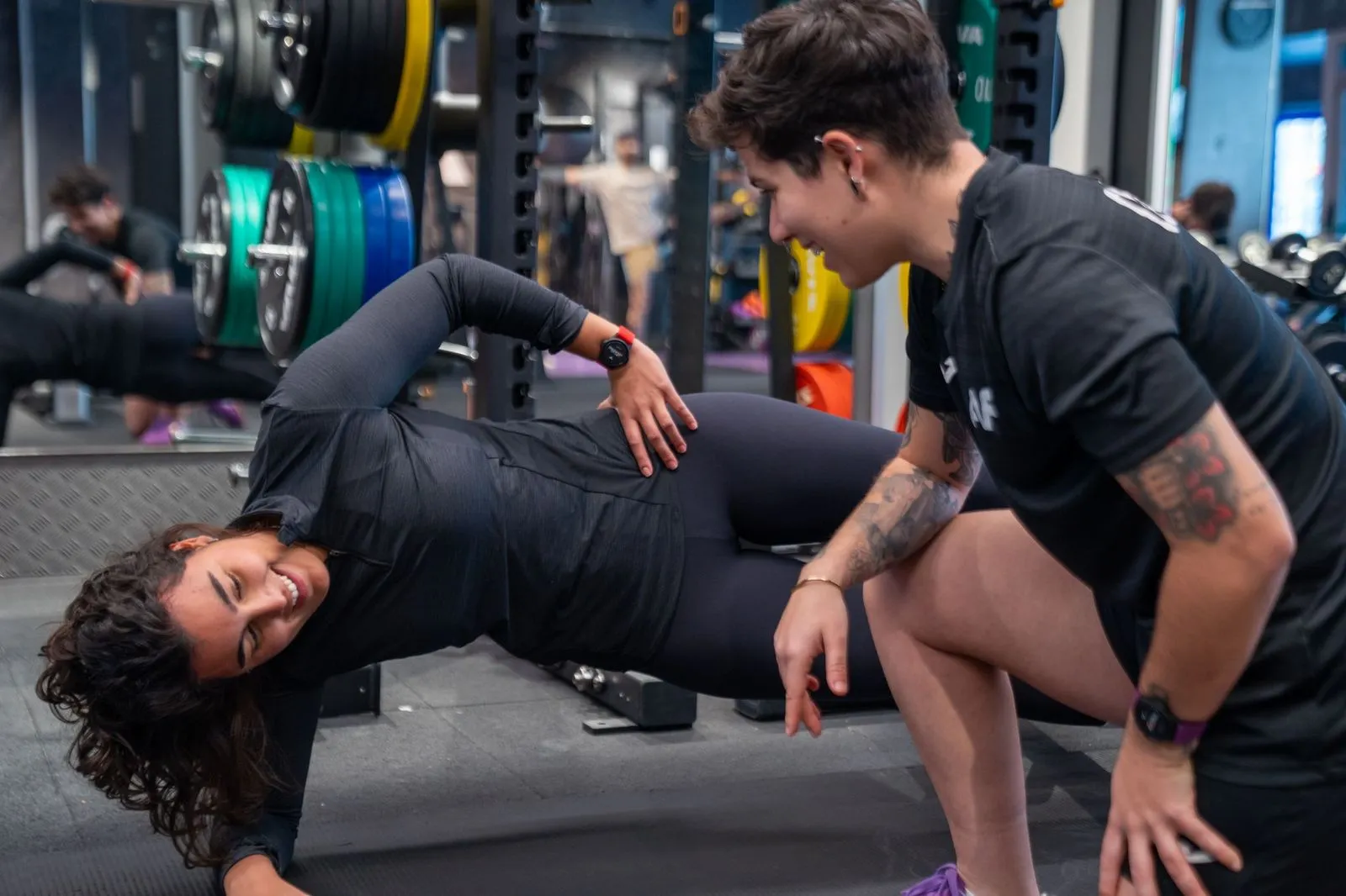 Una mujer que hace ejercicio de plancha lateral mientras es entrenadora la anima a ir al gimnasio con pesas en segundo plano.