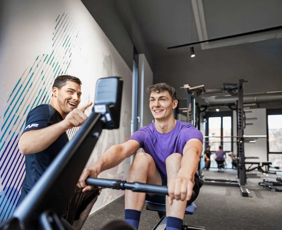 Ein Fitnesstrainer erklärt einem jungen Mann auf dem Rudergerät die Trainingswerte am Display.