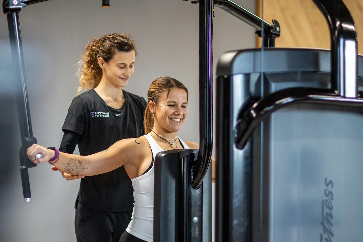 Eine Fitnesstrainerin betreut eine lächelnde Frau beim Training an der Schmetterlingsmaschine im Studio.